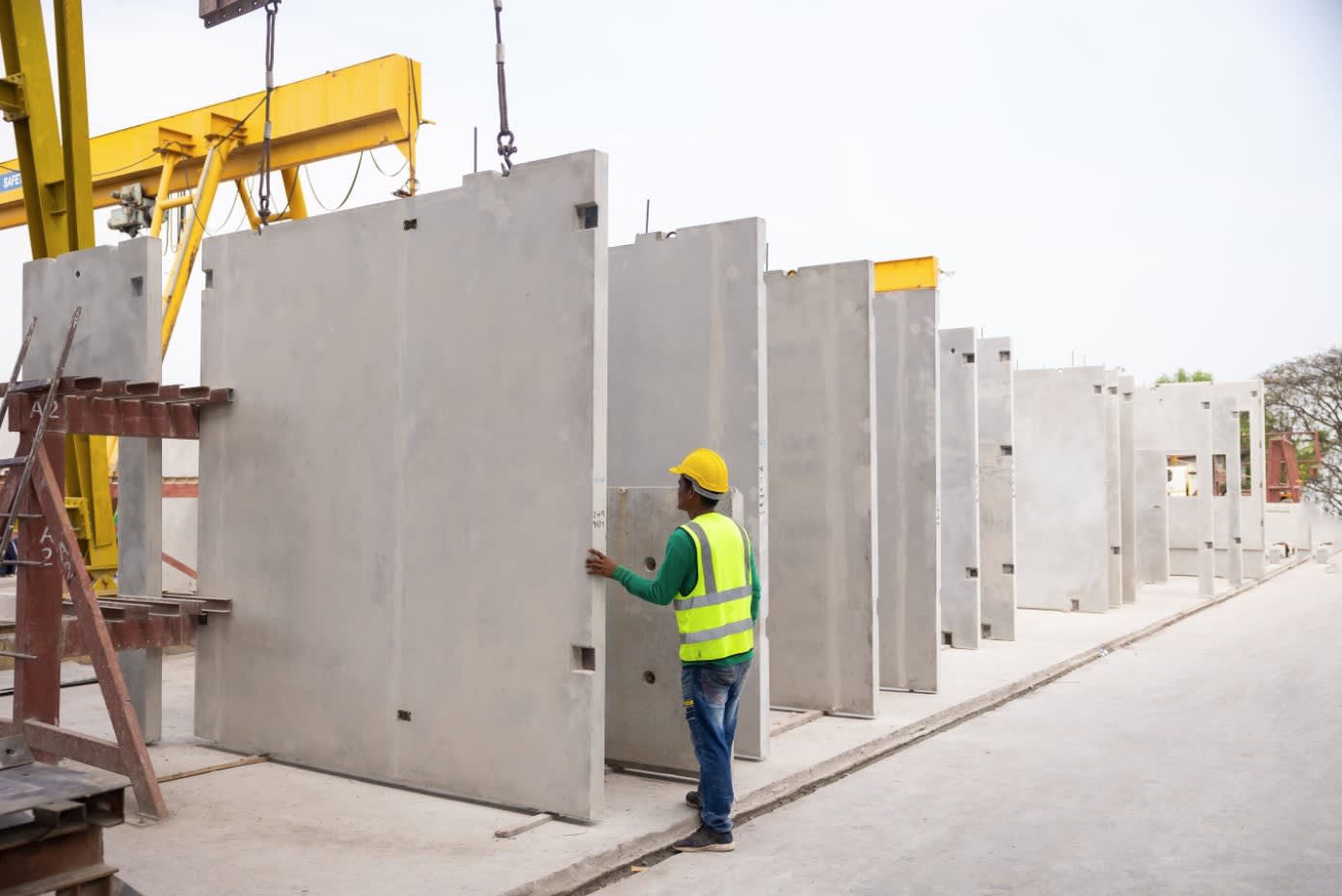 Operaio edile con casco giallo e giubbotto alta visibilità ispeziona un grande pannello prefabbricato in cemento armato, affiancato da altri pannelli simili e una gru a portale gialla in un cantiere.