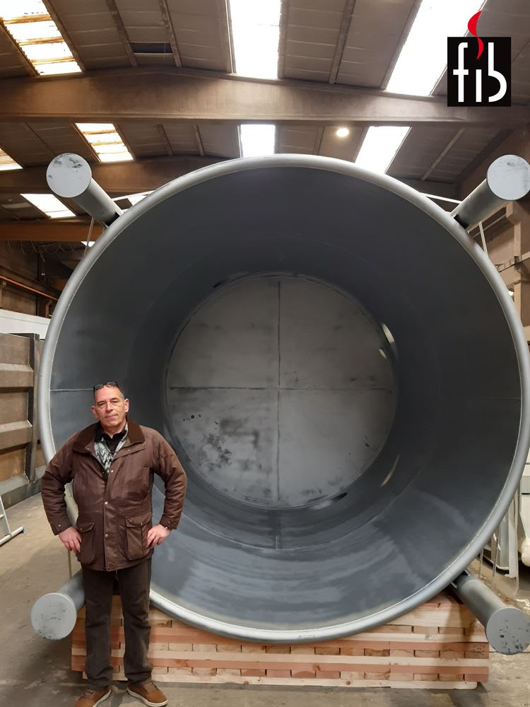 Man in a brown jacket standing beside a massive grey FIB industrial furnace tank, resting on wooden supports in a factory workshop.