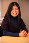 Smiling woman with long dark hair, wearing a dark blue turtleneck and a purple sweater, with hands clasped on a light-colored table.