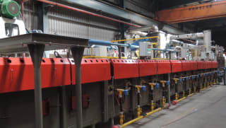 Red industrial continuous furnace or heat treatment line with overhead piping and control panels in a manufacturing facility.