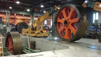 Yellow industrial crane positioned between massive orange and black grooved wheels in a heavy manufacturing facility, with other large industrial equipment in the background.