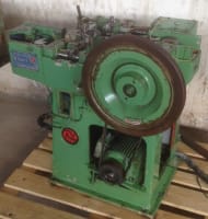 Green industrial nail making machine with a large brown flywheel, exposed mechanical components, and a visible motor, resting on a wooden pallet.