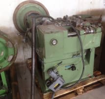 Green industrial production machine with a large top flywheel, pressure gauge, and pneumatic tool attachment, resting on a wooden pallet.