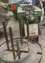 Industrial nail making machine processing wire fed from a large metal spool in a manufacturing facility.
