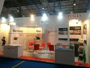 ITALIA trade show booth showcasing machinery and wire industries, featuring a reception desk, display cases, and a visitor seating area with red chairs.