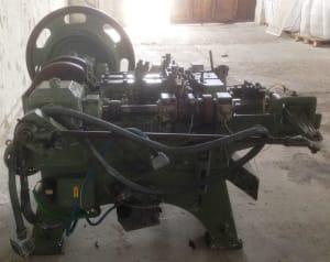 Green heavy-duty industrial manufacturing machine with a prominent flywheel, intricate gear assembly, and multi-station production line components.