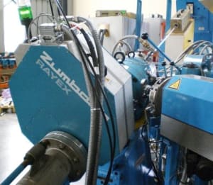 Blue and grey Zumbach RAYEX industrial measurement system on a production line, showing the hexagonal head and connected components.