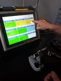 Mano che indica un monitor touch screen con un'interfaccia di gestione dati colorata, affiancato da un dispositivo di input specializzato e un calibro sul tavolo. Ambiente di lavoro digitale per controllo qualità o pianificazione produzione.