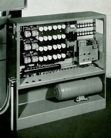 Open industrial electrical control panel with multiple rows of vintage relays, extensive wiring, terminal blocks, and a cylindrical capacitor on the bottom shelf.