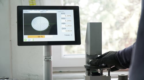 Operaio con guanto nero effettua misurazioni di precisione su un macchinario industriale, visualizzando i dati di controllo qualità (AVG, MAX, MIN) su un monitor digitale.