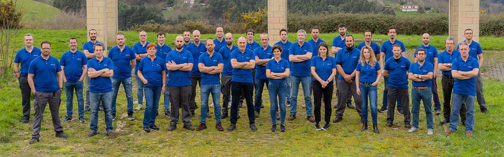 Company team portrait: A group of employees in blue polo shirts standing outdoors on a grassy field with hills and buildings in the background.