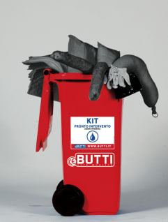 Red wheeled emergency spill kit bin, labeled 'KIT PRONTO INTERVENTO' and 'BUTTI', overflowing with gray absorbent pads, gloves, and spill response equipment.