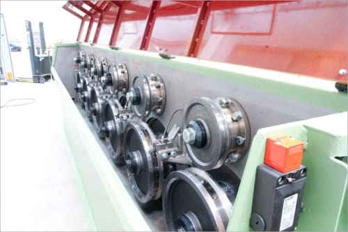 Macchina industriale per la lavorazione di materiali con una lunga serie di rulli metallici lucidi, telaio verde e copertura rossa, in un ambiente di produzione.