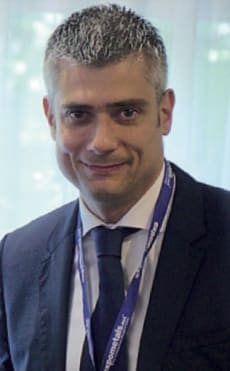 Uomo d'affari con capelli grigi, in completo e cravatta blu, sorride e indossa un badge da conferenza.