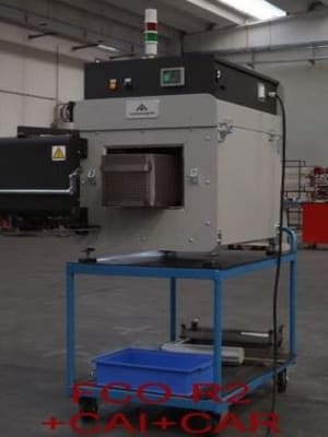 Grau-schwarze Industriemaschine auf einem blauen Rollwagen mit Bedienfeld, mehrfarbiger Signallampe und vorderer Öffnung in einer Fabrikhalle.