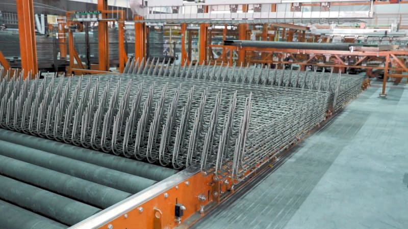 Stacks of prefabricated steel rebar cages moving on a roller conveyor system in a large industrial manufacturing plant.