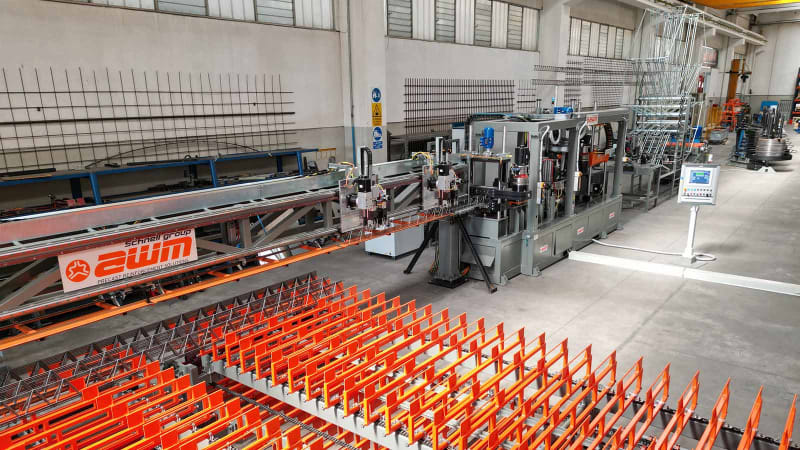 Automated AWM Schnell Group industrial machinery processing rebar for precast reinforcement solutions in a factory, featuring a long production line with welding stations and orange material handling racks.