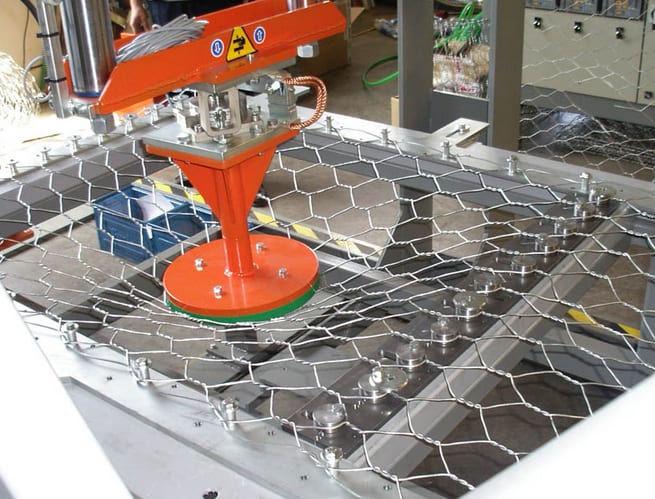 Automated arm with an orange circular tool pressing and deforming hexagonal wire mesh, likely for industrial material testing or manufacturing.