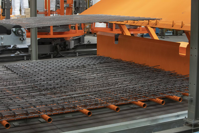 Industrial machine fabricating steel rebar mesh panels, showing the grid formation of metal bars with integrated orange conduits on a production line.