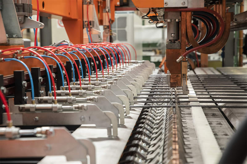 Máquina de soldadura industrial automatizada con múltiples conexiones hidráulicas/neumáticas rojas y azules, ensamblando una malla de refuerzo metálica.