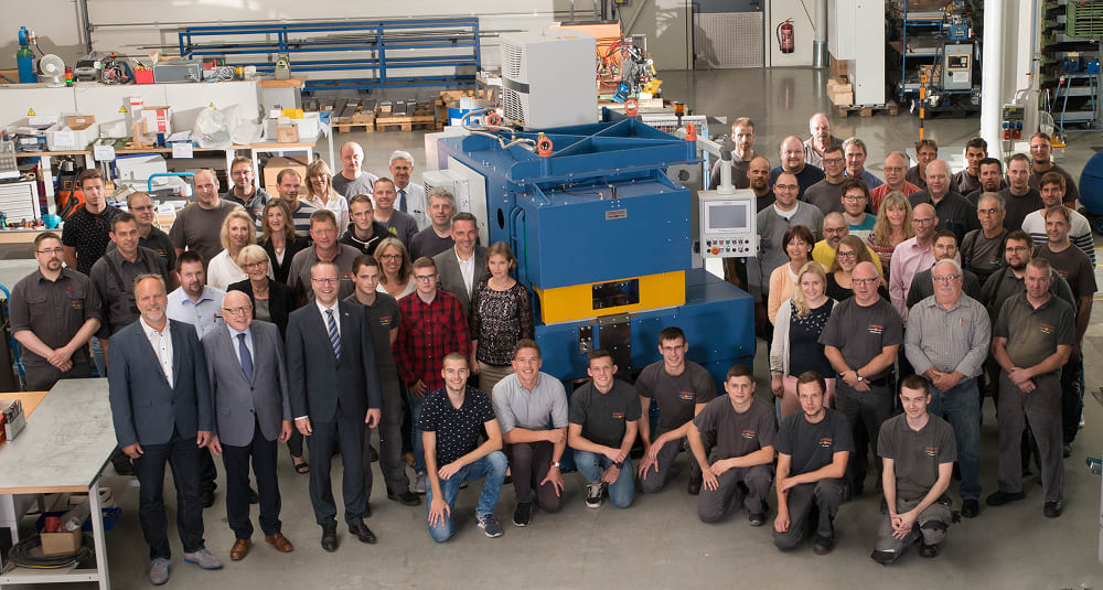 Gruppenfoto von engagierten Mitarbeitern eines Industrieunternehmens vor einer neuen blauen und gelben Produktionsmaschine in einer modernen Fertigungshalle.