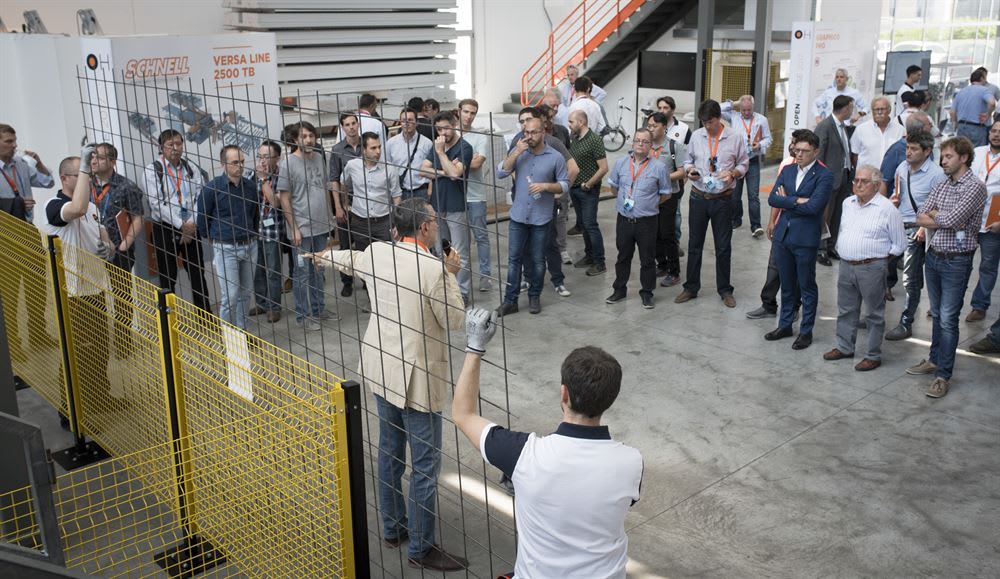 Présentation technique de la machine Schnell Versa Line 2500 TB à un groupe de professionnels, avec un expert expliquant les fonctionnalités derrière des barrières de sécurité jaunes.