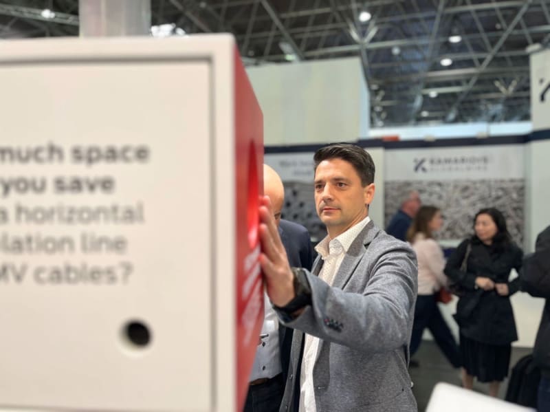 Professional man in a grey jacket interacting with an exhibition display providing information on MV cables, set against the backdrop of a busy trade show floor with K ZAMAKOVS branding.