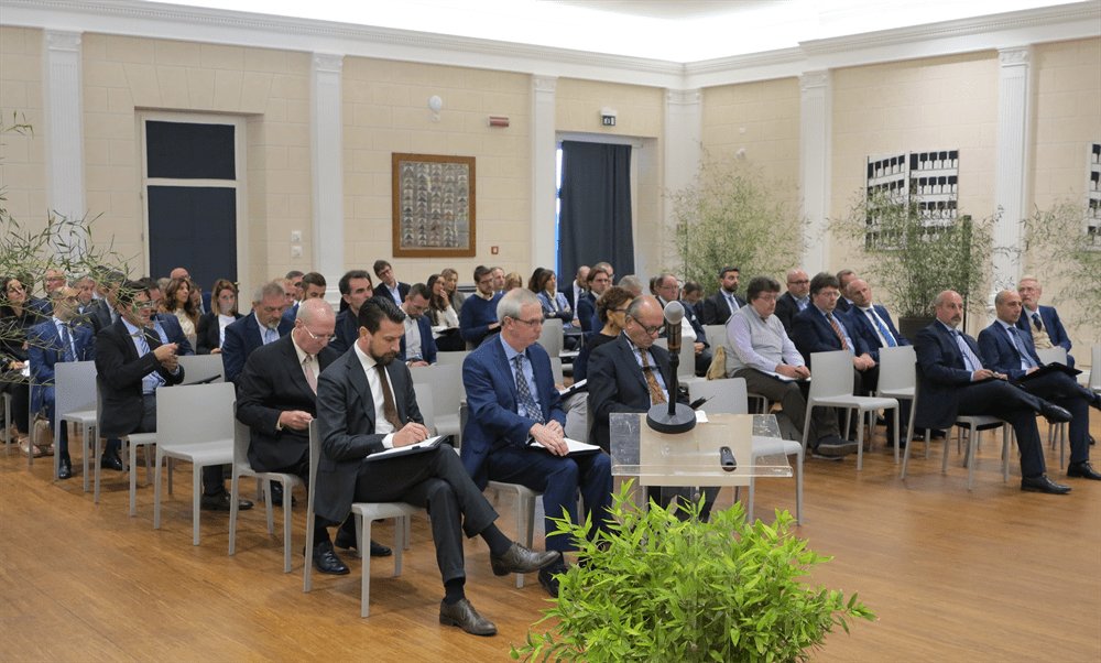 Professionals attending a business conference, seated in rows, with many taking notes in a well-lit meeting room.