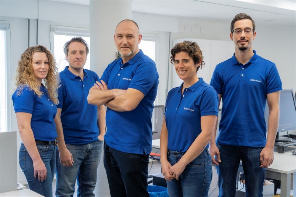 MEROLAM team photo featuring five smiling colleagues, three men and two women, wearing matching blue polo shirts in their office.