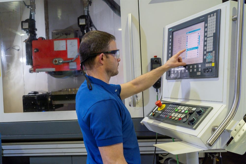 CNC machine operator wearing safety glasses, pointing at a Siemens control panel touchscreen display in a manufacturing facility.