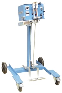 Light blue mobile industrial testing equipment with a top control unit, a clear vertical sample chamber, and foot pedal activation on a wheeled stand.