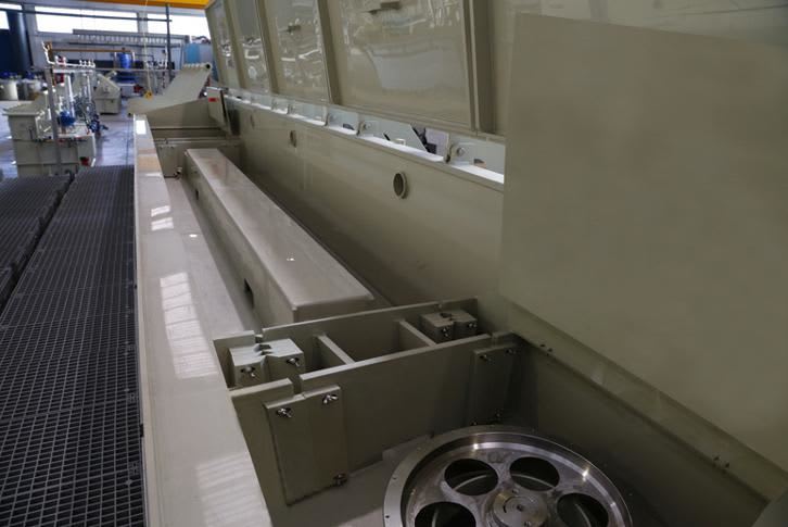 Interior view of a large light-colored industrial processing machine with an open section, revealing its internal support structures, a long central component, and a prominent metal spoked wheel, set in a factory environment with metal grated flooring.