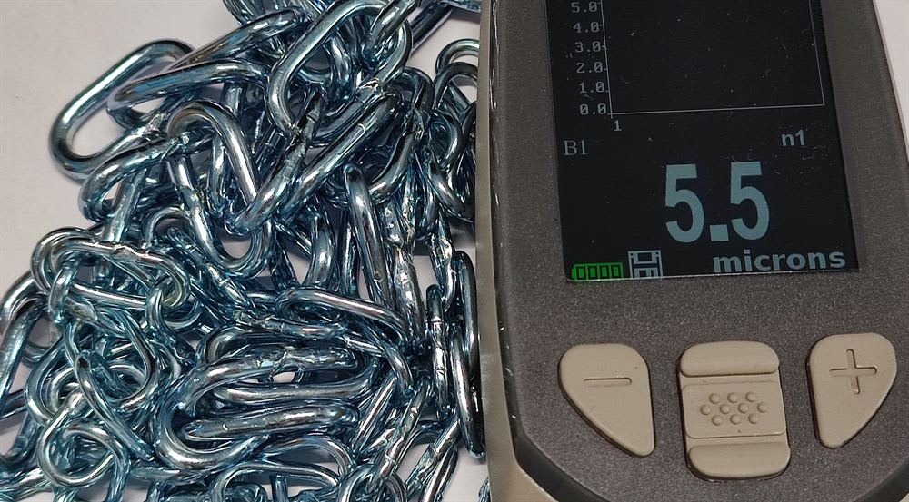 Galvanized chain links piled next to a digital coating thickness gauge displaying a measurement of 5.5 microns.