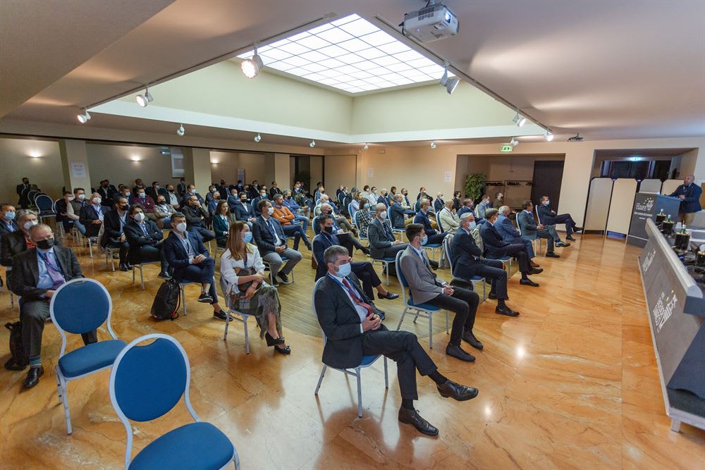 Conferenza professionale con pubblico seduto e distanziato in sala, molti indossano mascherine, e un relatore al podio.