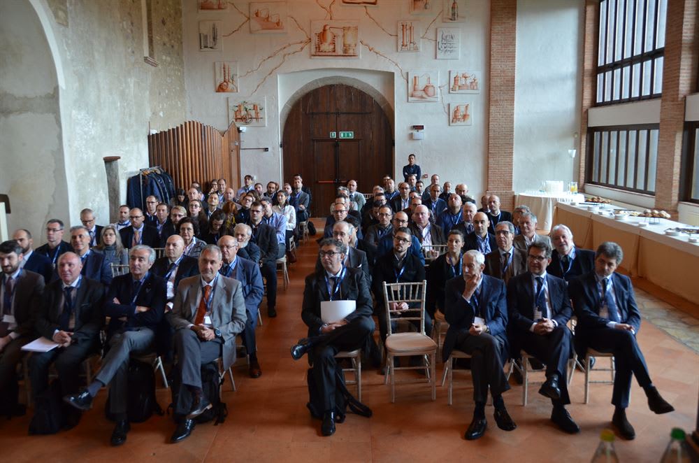 Pubblico di una conferenza professionale seduto in una sala storica con pareti decorate da illustrazioni a tema scientifico e un'area catering sullo sfondo.