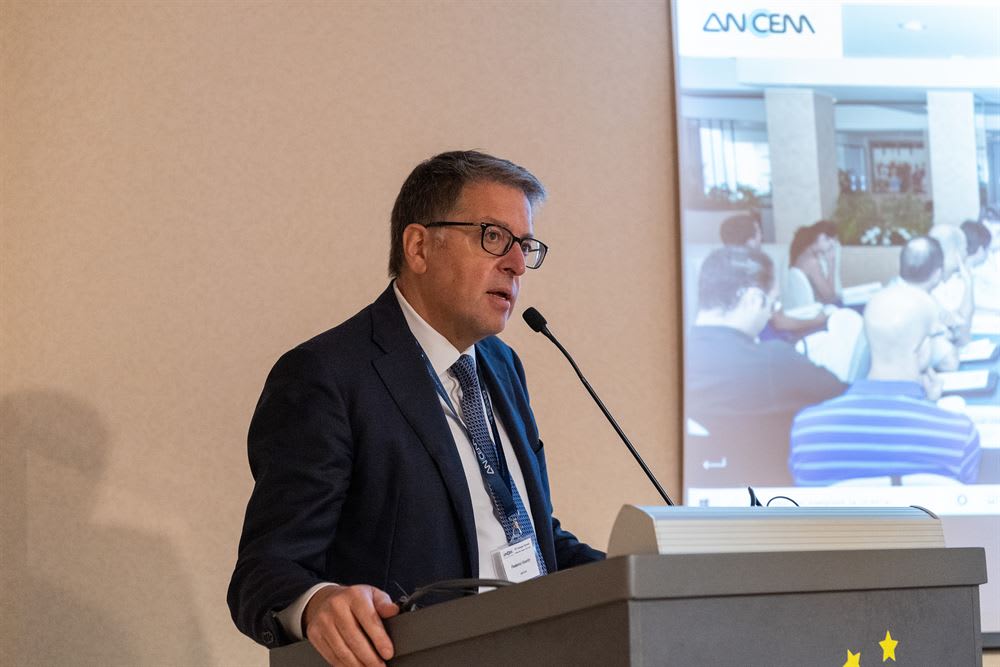 Relatore in completo e occhiali parla al microfono su un podio, con il logo ANCEM e un'immagine di partecipanti a una conferenza proiettati sullo sfondo.