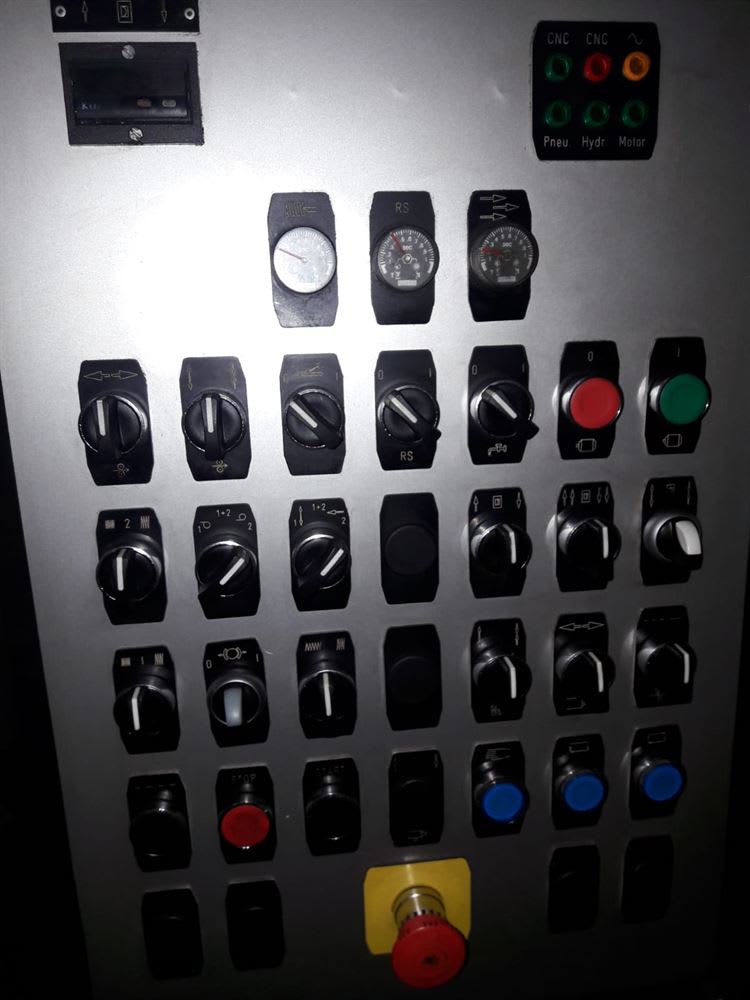 Industrial control panel featuring CNC indicators, a digital display, multiple rotary and toggle switches, various gauges, push buttons in red, green, and blue, and a prominent red emergency stop button.