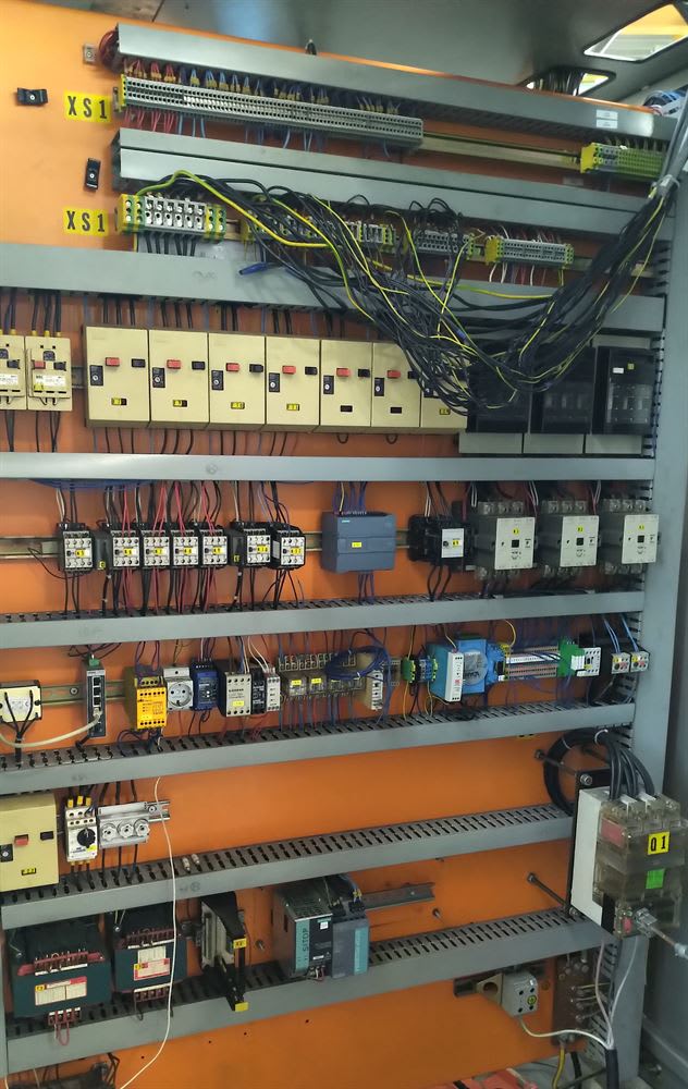 Open industrial electrical control panel with an orange backplate, showing multiple rows of DIN-rail mounted components, including circuit breakers, contactors, relays, programmable logic controllers (PLCs), and extensive electrical wiring.