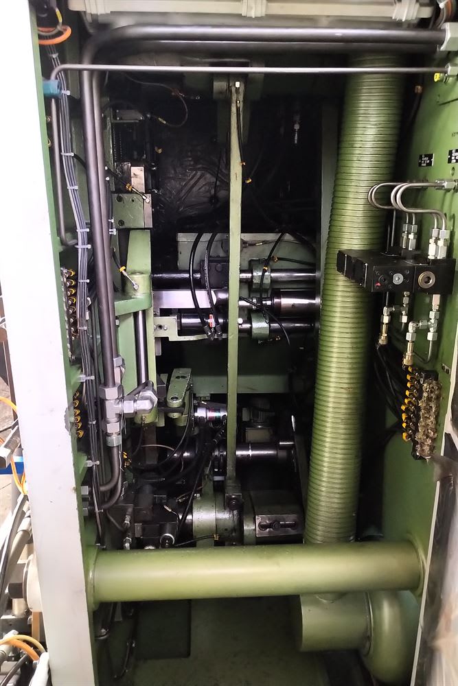 Internal view of a complex industrial machine, featuring multiple hydraulic cylinders, interconnected hoses, electrical wiring, and control valves. A large, ribbed green ventilation hose runs vertically on the right, all housed within the machine's robust green metal framework.