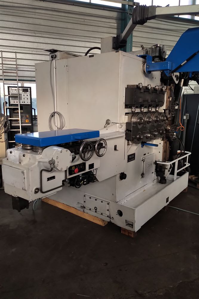 Large white industrial wire forming machine with numerous rollers, bending tools, control panels, and handwheels, set in a factory environment.