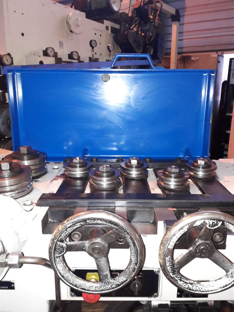 Industrial machine with a bright blue protective cover, a series of metal processing rollers, and two operator control handwheels.