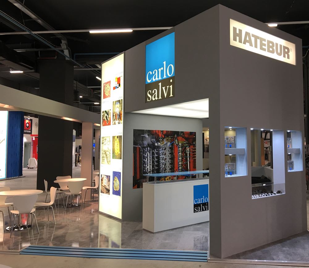 Two smiling business professionals, a woman and a man, stand behind a large rectangular yellow cake decorated with 'HATEBUR' and 'carlo salvi' logos, at a corporate event.
