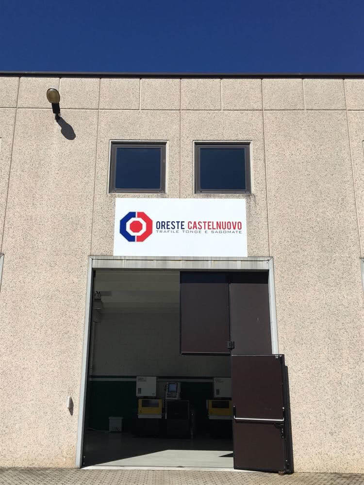 Exterior of ORESTE CASTELNUOVO building with 'TRAFILE TONDE E SAGOMATE' sign, an open industrial bay door revealing machinery, and a clear blue sky.