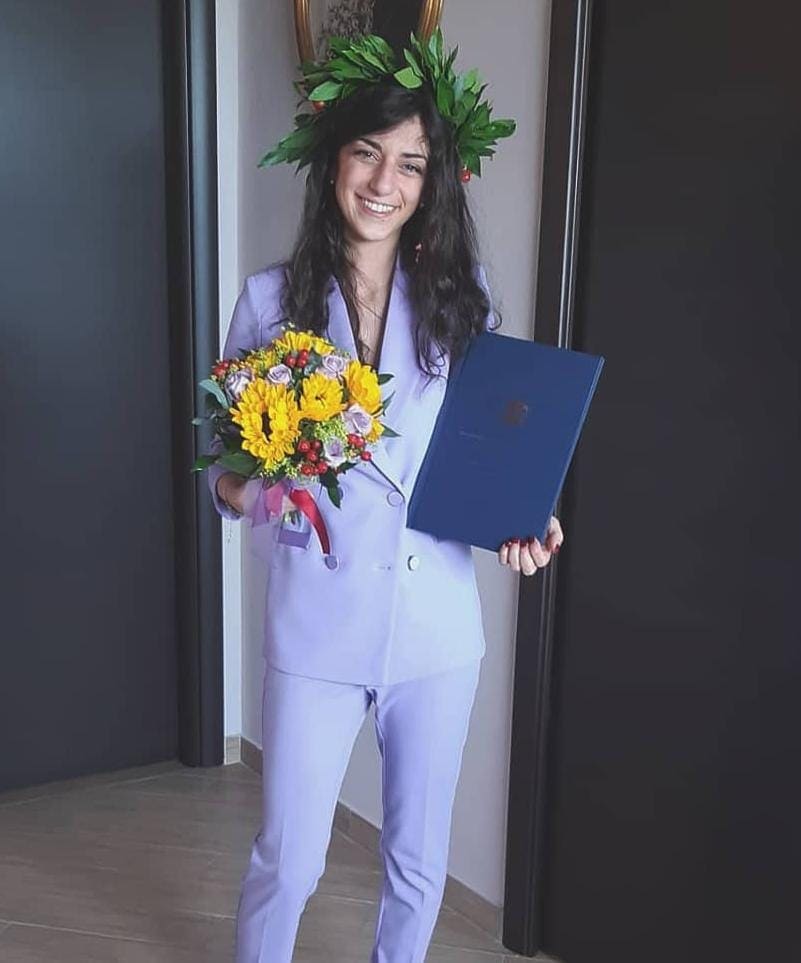 Smiling woman celebrating graduation, wearing a laurel wreath and a light purple pantsuit, holding a bouquet of sunflowers and a blue diploma folder.