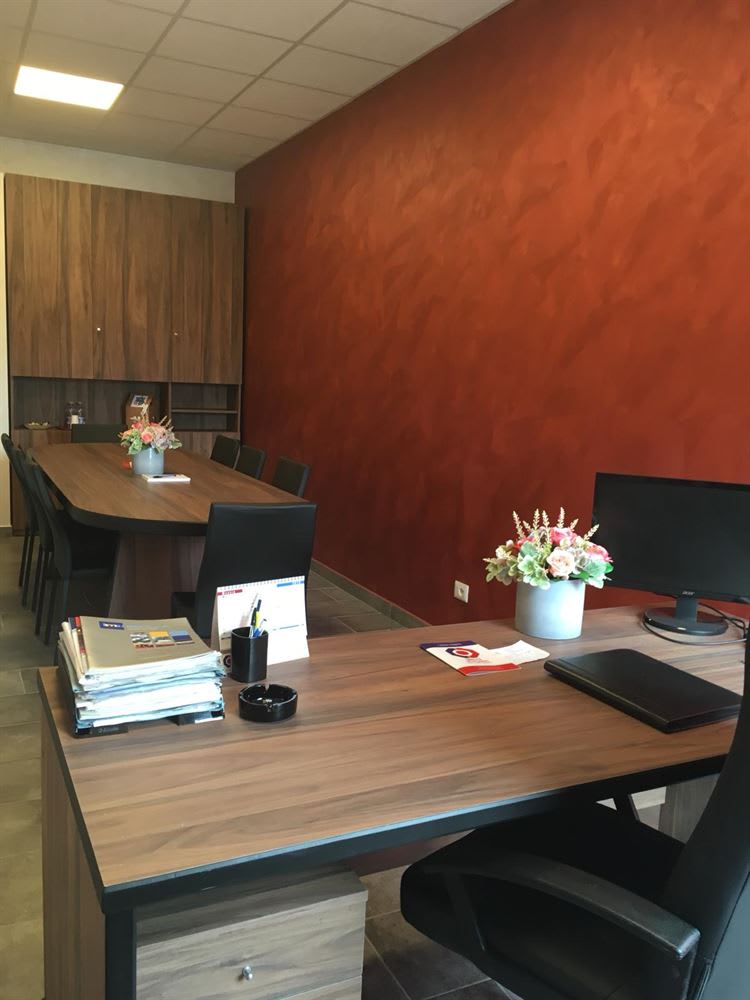 Modern office interior with a conference table, wooden storage cabinets, a vibrant red textured accent wall, and a desk with a computer monitor.