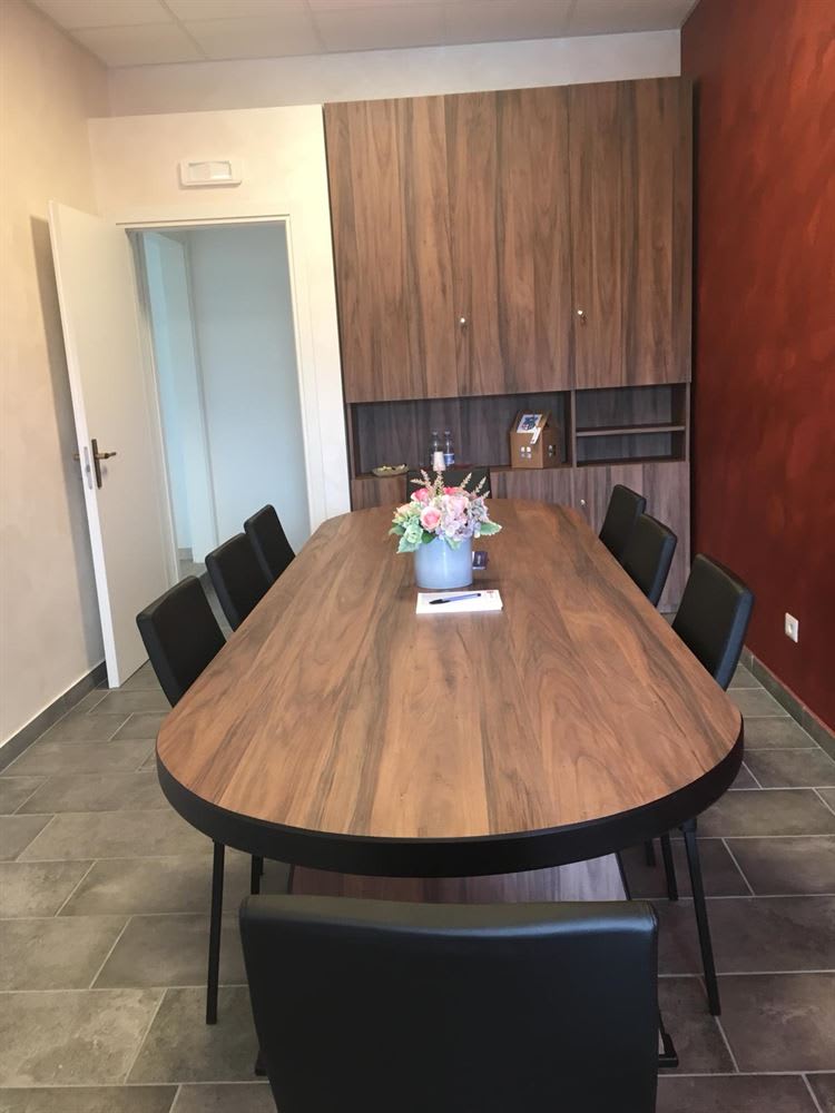 Modern conference room featuring an oval wooden meeting table with black chairs, a built-in wood storage cabinet, and a floral centerpiece. The room has grey tiled flooring and a red accent wall.