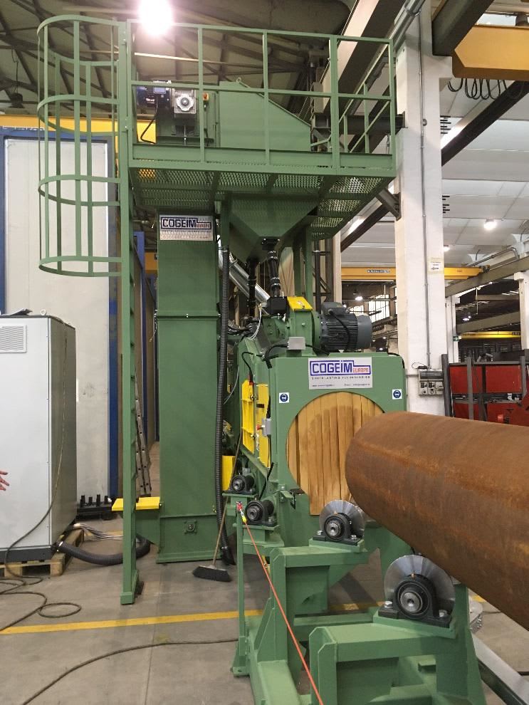 Impianto di granigliatura industriale COGEIM EUROPE per il trattamento superficiale di tubi, con struttura elevata, nastro trasportatore e rulliere di supporto, all'interno di un capannone di produzione.