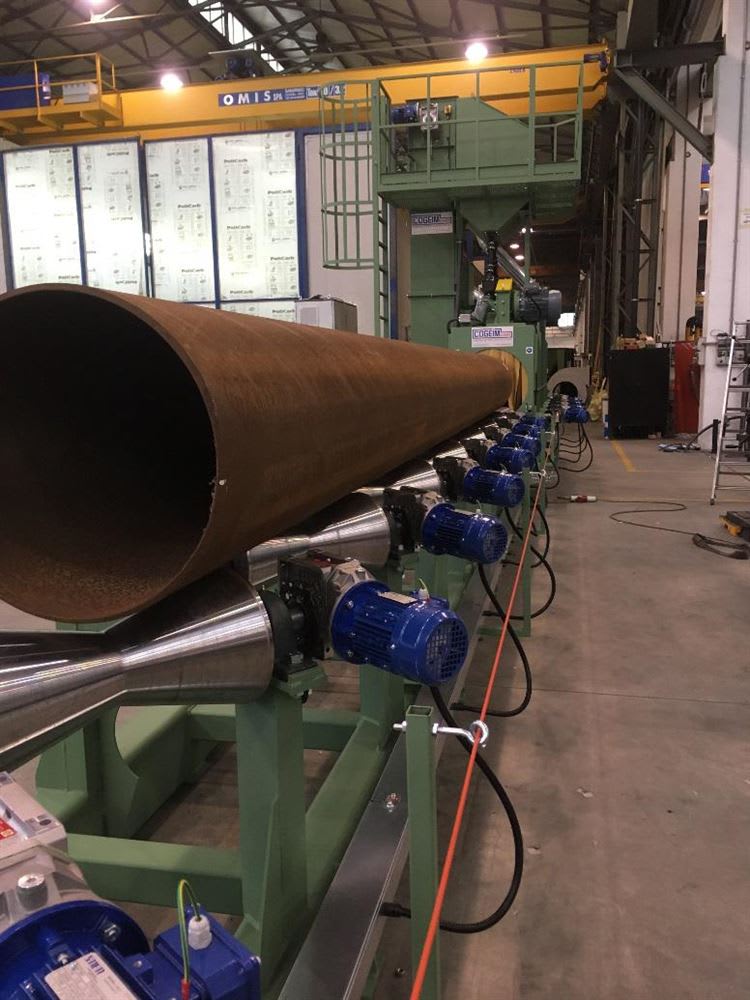 Grande tubo metallico arrugginito su un sistema di rulli motorizzati, in fase di movimentazione verso una macchina per la granigliatura COGIM all'interno di un impianto industriale.