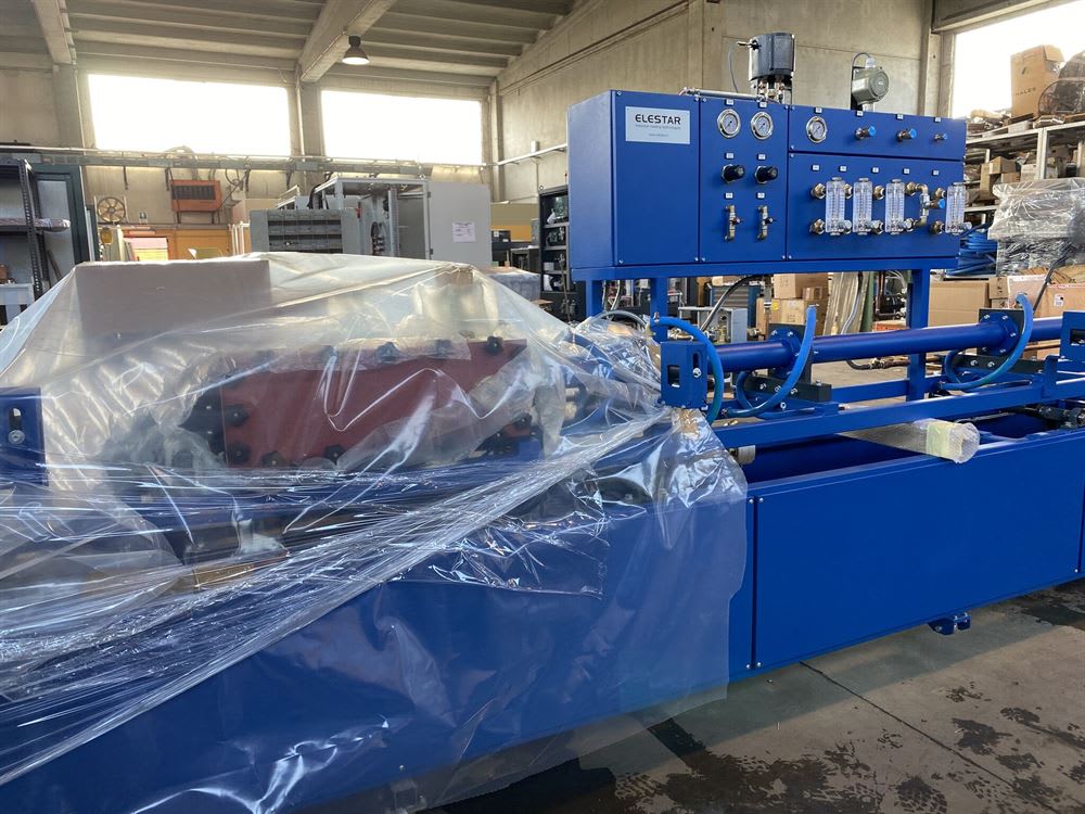 Macchinario industriale blu ELESTAR con pannello di controllo, manometri e flussimetri, parzialmente coperto da imballaggio protettivo in plastica, all'interno di un capannone.