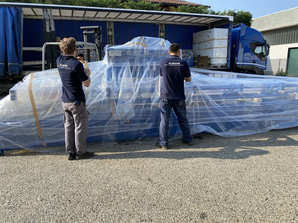 Due specialisti Elestar in divisa blu supervisionano la spedizione di un grande macchinario industriale blu, avvolto in pellicola protettiva, mentre un camion è in attesa per il trasporto.